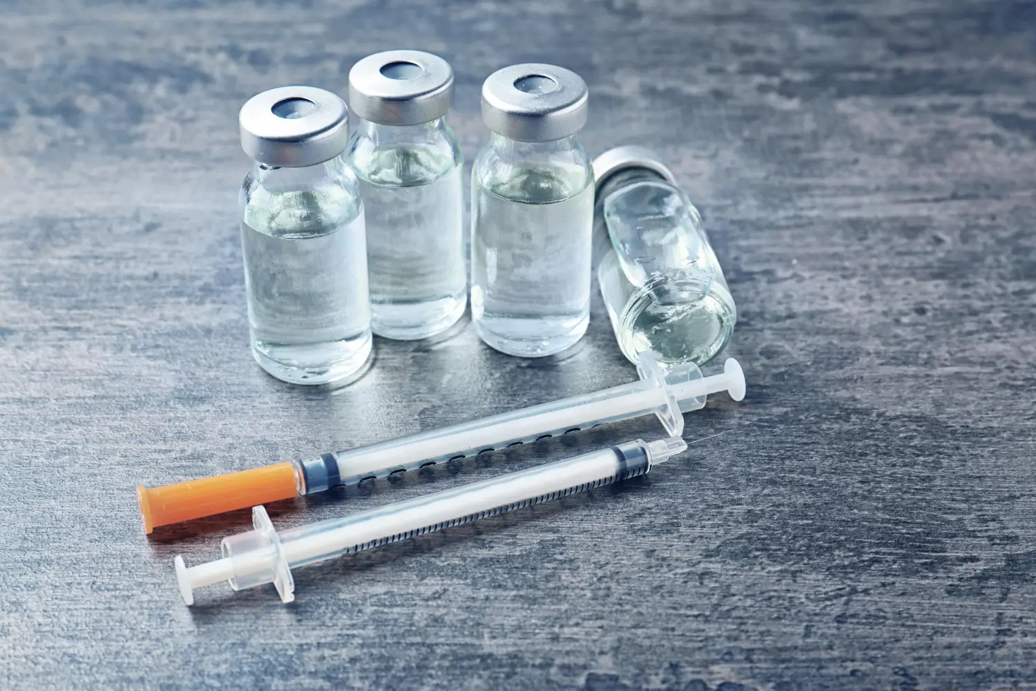 A close-up of syringes and vials on a gray background, representing injections, vaccinations, and medical treatment preparations.