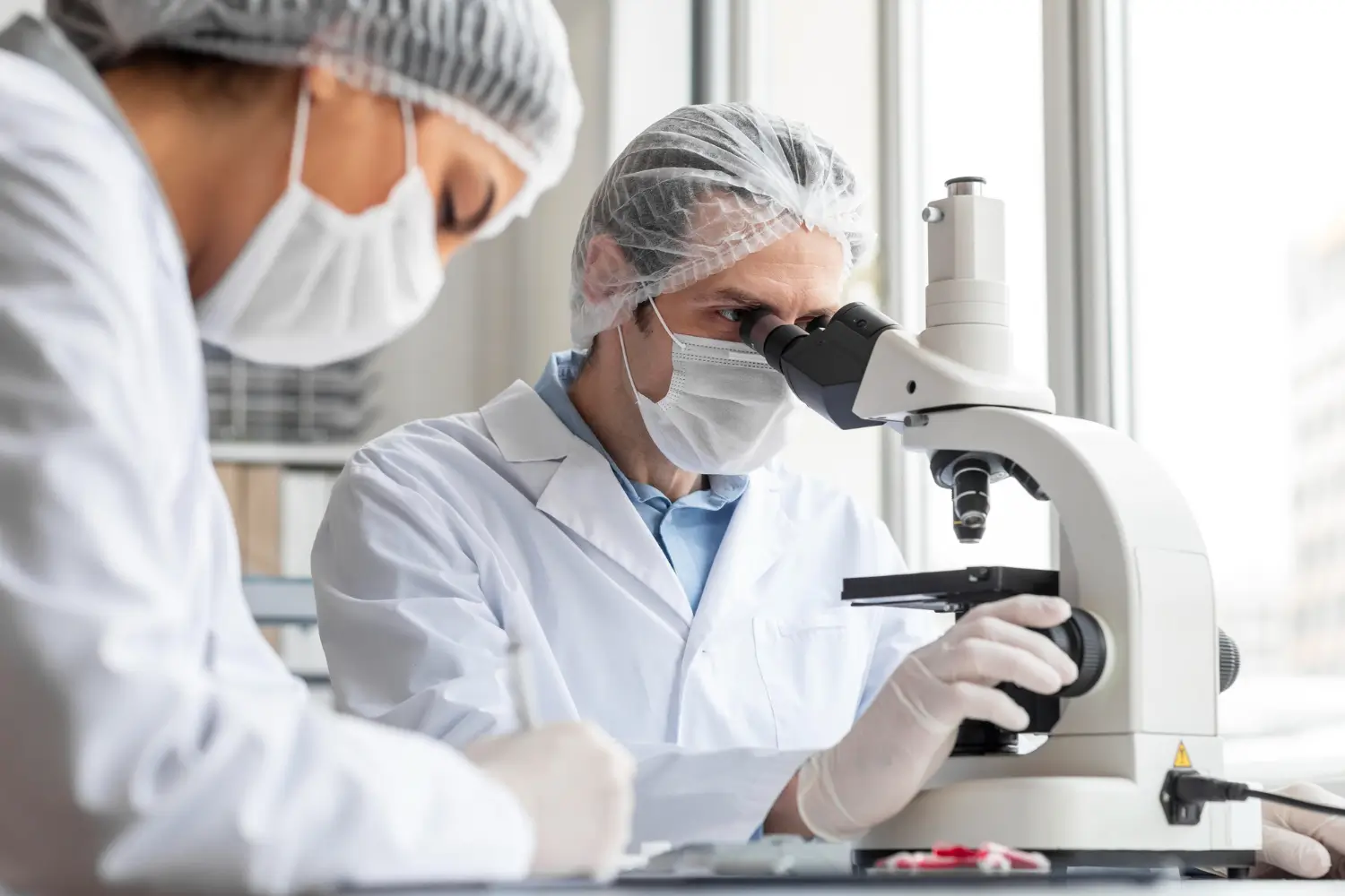 Scientists in PPE examine samples under a microscope for medical or biological analysis.